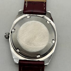 May include: A wristwatch with a circular silver-toned metal case and a smooth, brushed metal back. The watch features a small, ridged silver crown on the left side and is attached to a dark red leather strap with visible stitching and the text 18.