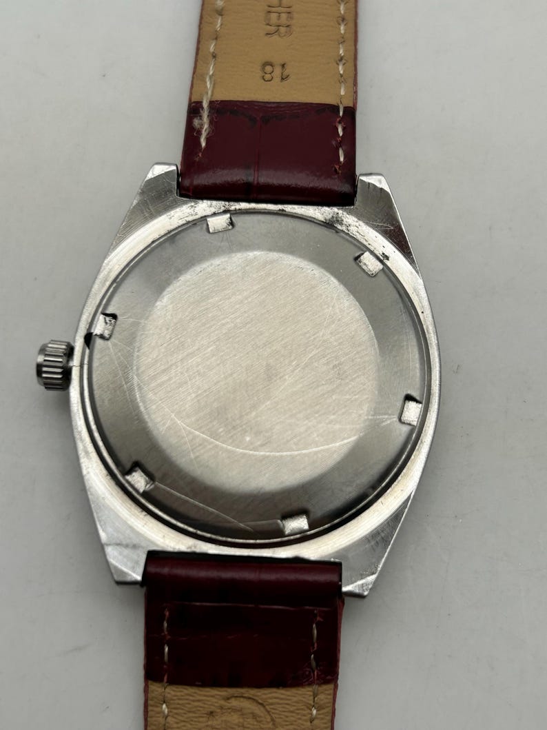 May include: A wristwatch with a circular silver-toned metal case and a smooth, brushed metal back. The watch features a small, ridged silver crown on the left side and is attached to a dark red leather strap with visible stitching and the text 18.