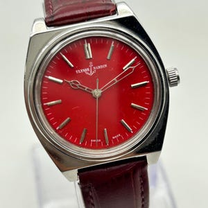 May include: A Ulysse Nardin wristwatch featuring a vibrant red dial with silver-tone hour markers and hands. The watch has a polished silver-tone metal case and a dark burgundy leather strap with a textured finish. The dial displays the brand name.