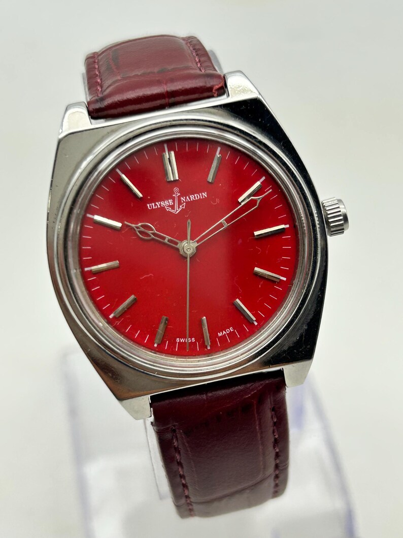 May include: A Ulysse Nardin wristwatch featuring a vibrant red dial with silver-tone hour markers and hands. The watch has a polished silver-tone metal case and a dark burgundy leather strap with a textured finish. The dial displays the brand name.