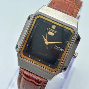 Könnte beinhalten: Vintage Seiko 5 Automatik-Uhr mit rechteckigem silberfarbenem Gehäuse und schwarzem Zifferblatt. Die Uhr hat goldfarbene Zeiger und Markierungen, eine Tages-Datumsanzeige und ein braunes Lederarmband.