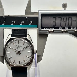 May include: A silver-toned Ulysse Nardin watch with a white face and black leather band is measured with a digital caliper. The watch face has silver hands and markers. The caliper displays a measurement of 37.40 mm.
