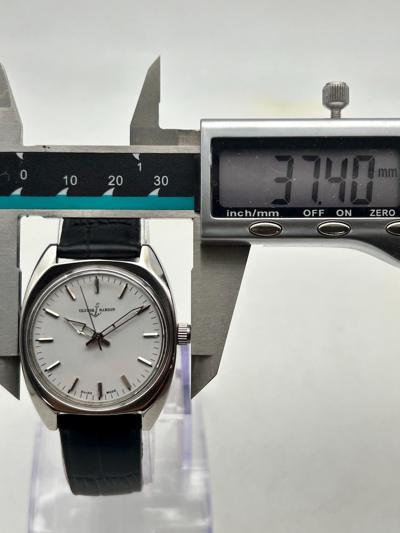 May include: A silver-toned Ulysse Nardin watch with a white face and black leather band is measured with a digital caliper. The watch face has silver hands and markers. The caliper displays a measurement of 37.40 mm.