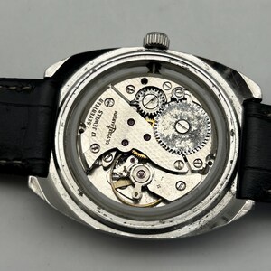 May include: Close-up of a vintage Ulysse Nardin watch with the back removed, revealing the intricate mechanical movement. The watch has a silver-toned case, a black leather strap, and the words "SEVENTEEN 17 JEWELS" are visible.