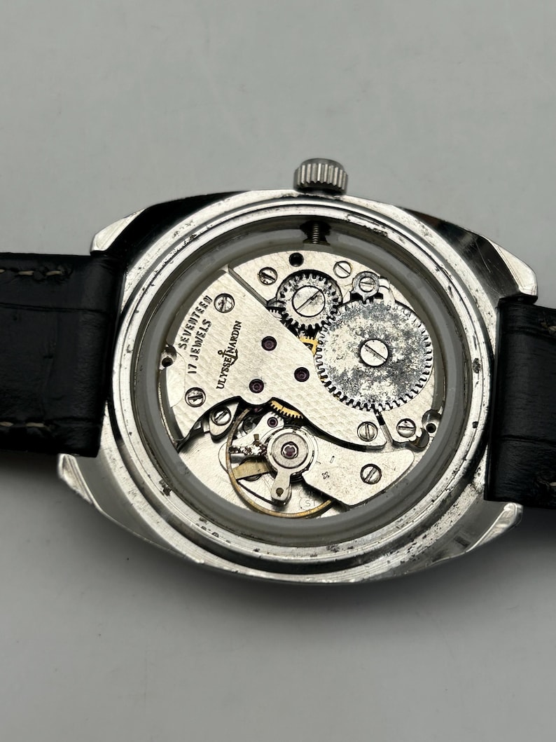 May include: Close-up of a vintage Ulysse Nardin watch with the back removed, revealing the intricate mechanical movement. The watch has a silver-toned case, a black leather strap, and the words "SEVENTEEN 17 JEWELS" are visible.