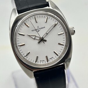 May include: A silver-toned Ulysse Nardin wristwatch with a white face and black leather band. The watch face features silver hands and markers, with the brand name and logo at the top. The words "SWISS MADE" are at the bottom.