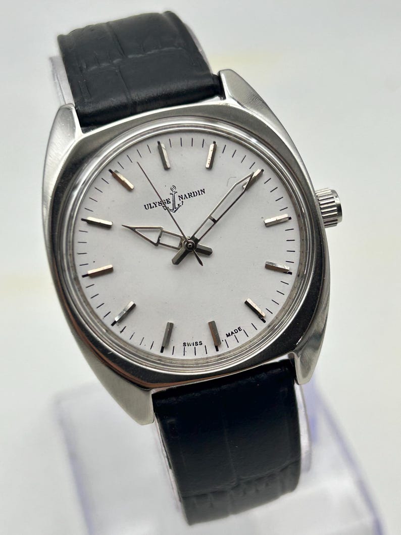 May include: A silver-toned Ulysse Nardin wristwatch with a white face and black leather band. The watch face features silver hands and markers, with the brand name and logo at the top. The words "SWISS MADE" are at the bottom.