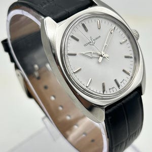 May include: A silver-toned Ulysse Nardin wristwatch with a white face and black leather band. The watch features silver hands and markers, and the band has a textured, crocodile-like pattern. The watch is displayed on a clear stand.