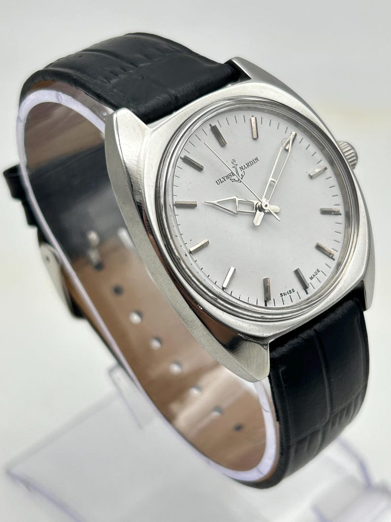 May include: A silver-toned Ulysse Nardin wristwatch with a white face and black leather band. The watch features silver hands and markers, and the band has a textured, crocodile-like pattern. The watch is displayed on a clear stand.