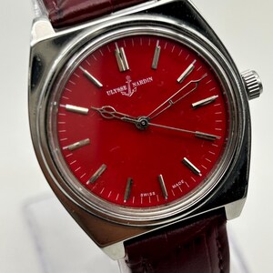 May include: A Ulysse Nardin wristwatch featuring a vibrant red dial with silver-tone hour markers and hands. The watch has a polished silver-tone metal case and a dark burgundy leather strap with a textured, embossed pattern.