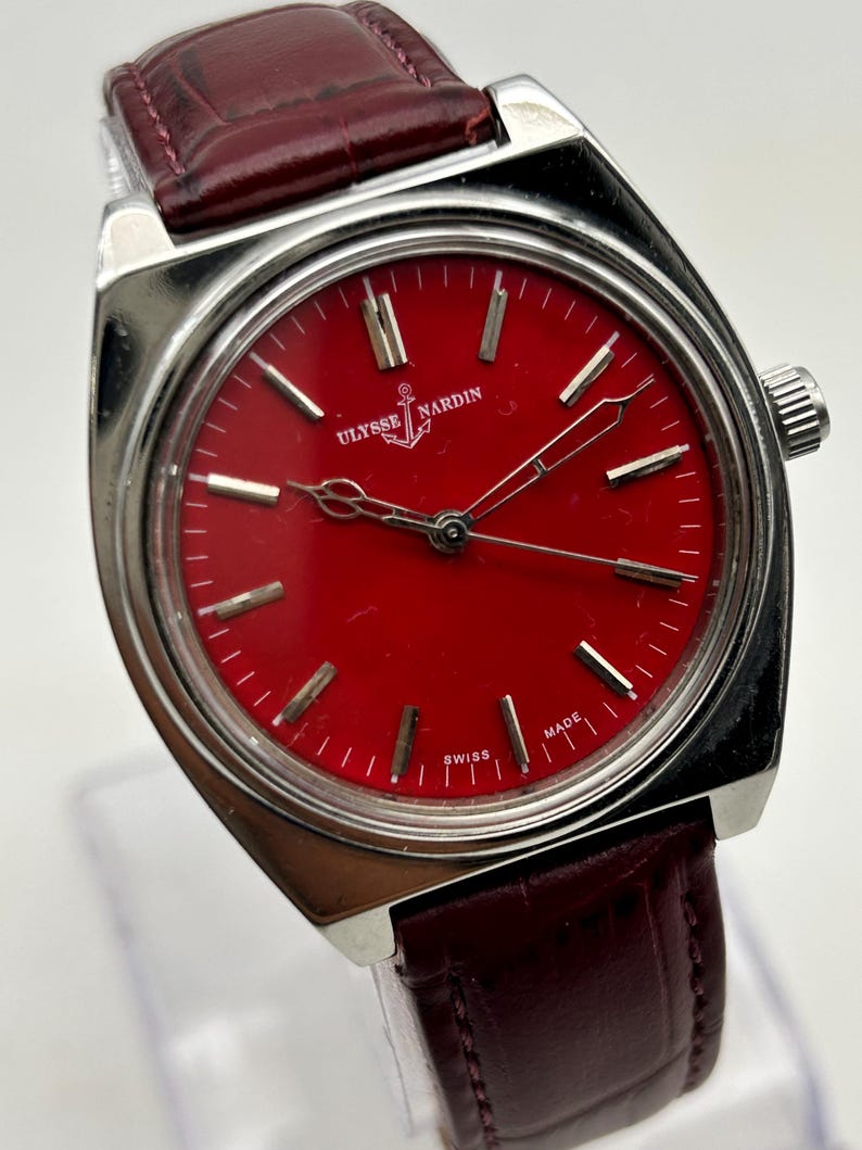 May include: A Ulysse Nardin wristwatch featuring a vibrant red dial with silver-tone hour markers and hands. The watch has a polished silver-tone metal case and a dark burgundy leather strap with a textured, embossed pattern.