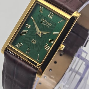 May include: A rectangular gold-toned Seiko Quartz watch with a green face and gold Roman numerals. The watch has a brown leather band. The watch face has a textured pattern and the Seiko logo.