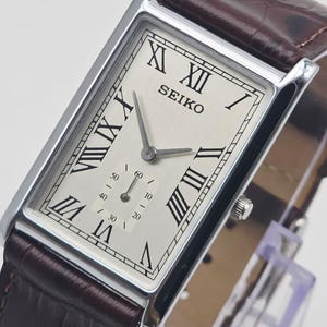 May include: A rectangular Seiko wristwatch with a silver face, black Roman numerals, and a small seconds dial. The watch has a brown leather strap with a crocodile pattern and a silver case.