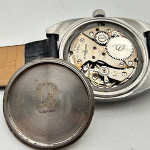 May include: A vintage Ulysse Nardin wristwatch with a black leather strap. The watch case is open, revealing the intricate mechanical movement with gears and jewels. The back of the watch case is detached, showing engraved text and a logo.