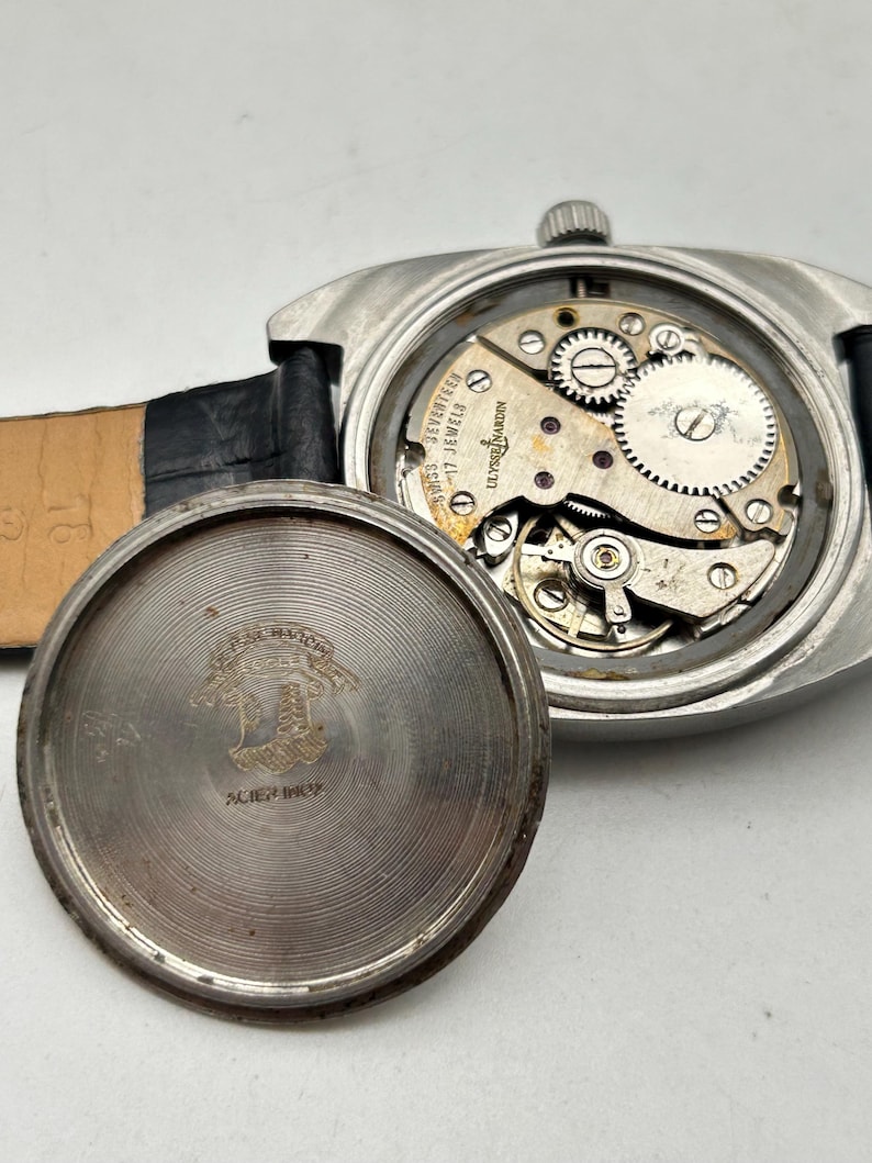 May include: A vintage Ulysse Nardin wristwatch with a black leather strap. The watch case is open, revealing the intricate mechanical movement with gears and jewels. The back of the watch case is detached, showing engraved text and a logo.