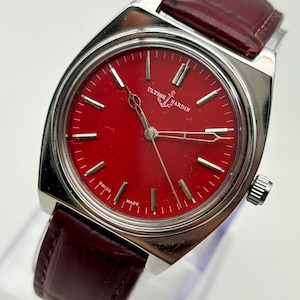 May include: A Ulysse Nardin wristwatch featuring a vibrant red dial with silver-tone hour markers and hands. The watch has a polished silver-tone metal case and a dark burgundy leather strap with a textured finish. The dial displays the brand name.