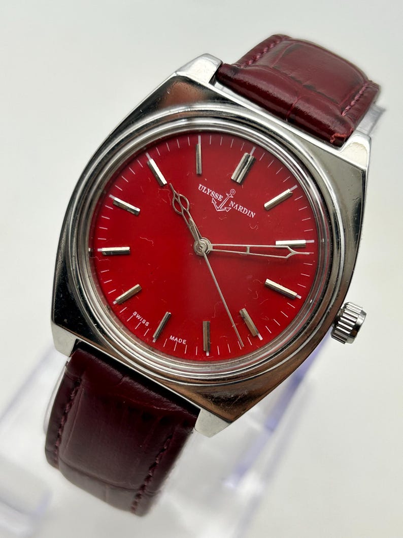 May include: A Ulysse Nardin wristwatch featuring a vibrant red dial with silver-tone hour markers and hands. The watch has a polished silver-tone metal case and a dark burgundy leather strap with a textured finish. The dial displays the brand name.
