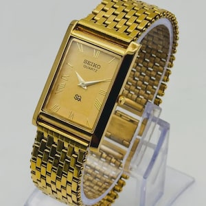 May include: A gold-toned Seiko Quartz watch with a rectangular face and a matching gold band. The watch face displays Roman numerals and the Seiko Quartz logo. The watch is displayed on a clear plastic stand.