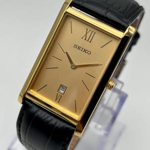 Peut inclure: Une montre Seiko dorée avec un cadran rectangulaire et un bracelet en cuir noir. Le cadran affiche le chiffre XII et le nom de la marque. La date est affichée comme le chiffre 20. La montre a une lunette dorée et un bracelet en cuir noir.