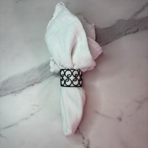 May include: A white fabric napkin folded and secured with a black metal napkin ring. The napkin ring features a repeating openwork scroll pattern made of thin wire. The fabric of the napkin has a visible textured weave and soft, draped folds.