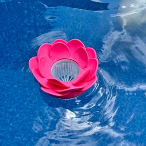 May include: A bright pink floating pool filter shaped like a lotus flower. The filter has a white mesh basket in the center and is surrounded by blue water. The filter is designed to collect debris.
