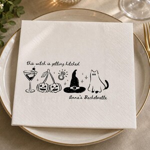 May include: A square white paper napkin featuring black ink illustrations of a martini glass with a ghost, two jack-o'-lantern cherries, a diamond ring, a witch hat, and a ghost cat. Text reads: this witch is getting hitched Anna's Bachelorette.