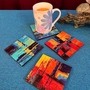 May include: A white mug with floral designs sits on a colorful square coaster. Three additional coasters with abstract art in shades of blue, yellow, orange, and red are also visible. The coasters appear to be made of wood.