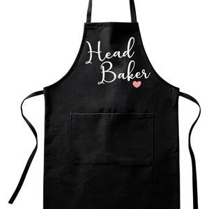 May include: Black apron with the words "Head Baker" in white script, a small pink heart, and a front pocket. The apron has a textured surface and black ties at the waist and neck.