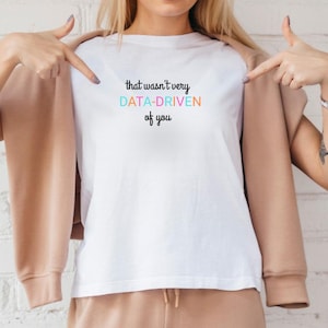 Puede incluir: Camiseta blanca con la frase "that wasn't very DATA-DRIVEN of you" en texto negro y colorido. Una persona señala la camiseta.