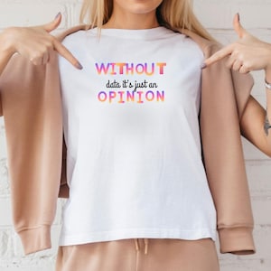 Peut inclure: T-shirt blanc avec le texte "WITHOUT data it's just an OPINION" en lettres dégradées colorées. Le t-shirt est montré par deux mains. La personne porte une veste et un pantalon beiges.