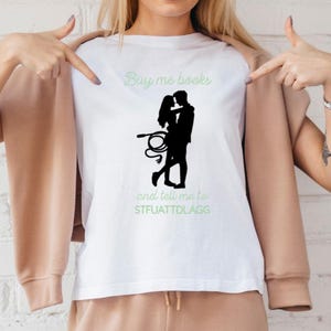 Könnte beinhalten: Weißes T-Shirt mit einer schwarzen Silhouette eines Paares, einer Peitsche und dem Text "Buy me books and tell me to STFUATTDLAGG" in Grün und Schwarz. Eine Person zeigt auf das T-Shirt.