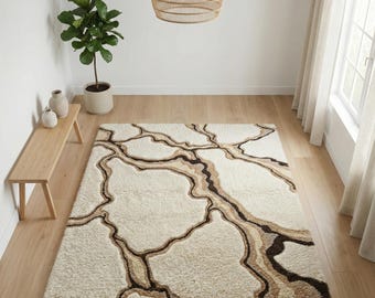 Puede incluir: Una alfombra rectangular de color crema con un diseño abstracto marrón, beige y crema. La alfombra tiene una textura peluda y está colocada sobre un suelo de madera clara. La habitación también cuenta con una planta, un banco y una lámpara tejida.