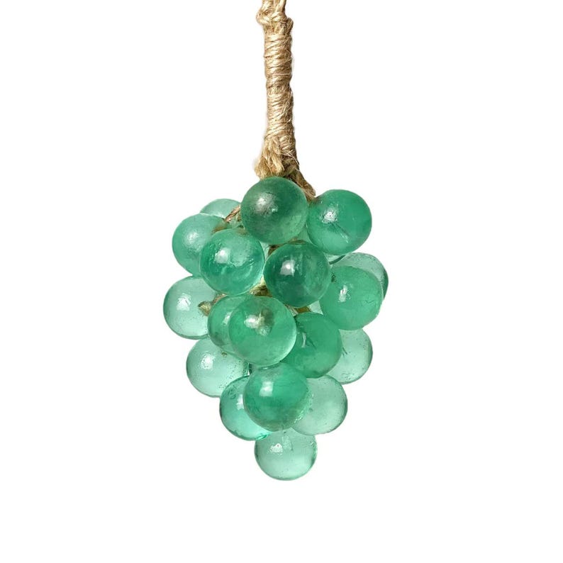 May include: A cluster of green glass spheres resembling grapes, suspended from a natural fiber cord. The spheres vary slightly in size and shade, creating a textured, organic appearance. The background is plain white.
