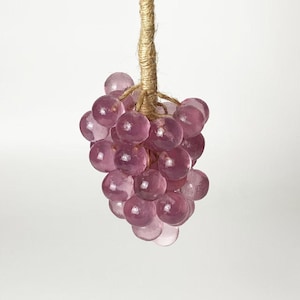 May include: A cluster of translucent, purple glass grapes hangs from a natural twine stem. The grapes are round and vary in size, with a slight variation in color. The twine stem is tightly wound, and the background is a plain white.