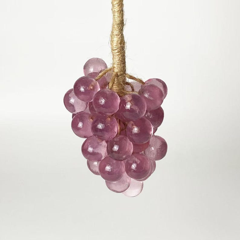May include: A cluster of translucent, purple glass grapes hangs from a natural twine stem. The grapes are round and vary in size, with a slight variation in color. The twine stem is tightly wound, and the background is a plain white.