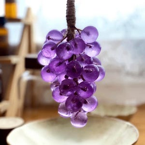 May include: A cluster of translucent purple grapes, suspended by a brown, textured stem. The grapes are round and appear to be made of glass or a similar material. The background is blurred, suggesting a shallow depth of field.