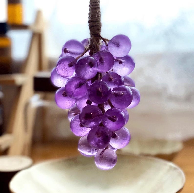 May include: A cluster of translucent purple grapes, suspended by a brown, textured stem. The grapes are round and appear to be made of glass or a similar material. The background is blurred, suggesting a shallow depth of field.