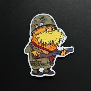 Może przedstawiać: Naklejka przedstawiająca postać z kreskówki z żółtym futrem i dużymi żółtymi wąsami, ubraną w wojskowy hełm i mundur, trzymającą karabin. Na hełmie widnieje napis "DEATH FROM ABOVE". Postać ma na sobie czerwoną chustę.