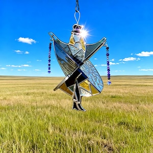 May include: Stained glass suncatcher in the shape of a figure with outstretched arms, featuring geometric shapes in blue, yellow, black, and clear glass. The suncatcher is suspended by a black cord and adorned with blue bead strands. Set against a backdrop of a blue sky and a grassy field.