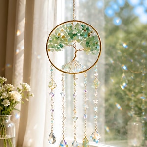 Puede incluir: Un atrapasol colgante con un marco circular dorado que contiene una escultura de árbol hecha con alambre y fragmentos de piedra de color verde claro y translúcido. Cinco cadenas colgantes incluyen cuentas de cristal, estrellas y gotas de vidrio.