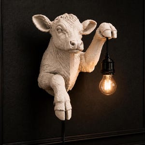 May include: A wall-mounted light fixture featuring a sculpted white calf head and front leg. The calf is depicted holding a black pendant light socket with a glowing, clear glass light bulb suspended from its hoof, creating a functional lamp.