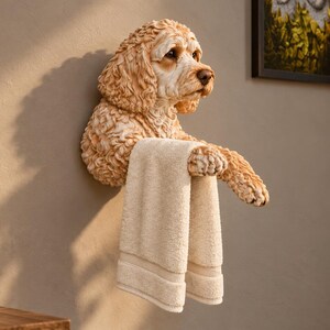 Puede incluir: Una escultura de pared de un perro de color marrón claro y pelo rizado con las patas delanteras extendidas hacia adelante. Las patas del perro están posicionadas para sostener una toalla doblada de color crema, funcionando como toallero.