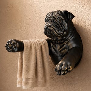 Op de afbeelding: Een donkerbronskleurige wandhanddoekhouder in de vorm van de kop en voorpoten van een buldog. Het beeldhouwwerk toont gedetailleerde gezichtsrimpels en poten, met een beige handdoek gedrapeerd over een van de uitgestrekte voorpoten.