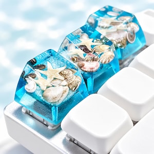 Puede incluir: Dos teclas mecánicas personalizadas hechas de resina azul translúcida, cada una con estrellas de mar blancas y varias conchas pequeñas incrustadas en una base arenosa, instaladas en un teclado blanco junto a teclas blancas.