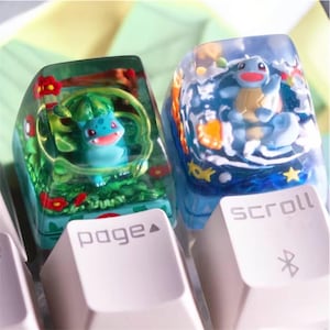 Puede incluir: Dos teclas de teclado personalizadas con diseños de personajes de dibujos animados. Una tecla presenta un personaje azul y verde en un entorno verde con flores rojas. La otra tecla tiene un personaje azul en una escena acuática. Teclas de teclado blancas.