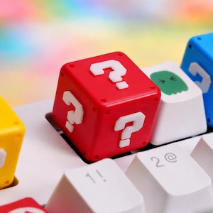 Peut inclure: Un capuchon de touche rouge en forme de cube avec des points d'interrogation blancs sur chaque face, placé sur un clavier blanc. D'autres capuchons de touches sont visibles, dont un fantôme vert et des cubes bleus et jaunes. Les capuchons de touches sont conçus pour ressembler à des éléments de jeu vidéo.