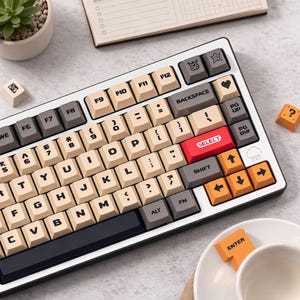 Könnte beinhalten: Eine mechanische Tastatur mit weißem Rahmen, schwarzer Basis und beigefarbenen Tastenkappen. Die Tasten haben schwarze Buchstaben und enthalten orange, rote und graue Akzenttasten. Die Tastatur hat eine Vielzahl von Symbolen und Icons auf den Tasten, darunter ein Herz, ein Stern und eine Wolke.