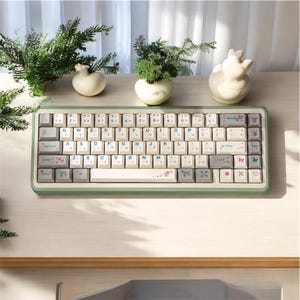 May include: A mint green and cream-colored mechanical keyboard with Japanese characters and floral accents. The keyboard has a standard layout with a white space bar and decorative keycaps. The keyboard is surrounded by small decorative items and greenery.