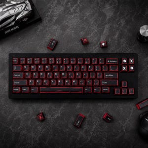 Puede incluir: Teclado mecánico negro con teclas retroiluminadas en rojo. El teclado tiene un diseño japonés con caracteres en inglés. Varias teclas rojas están esparcidas alrededor del teclado. El teclado está sobre una superficie oscura.