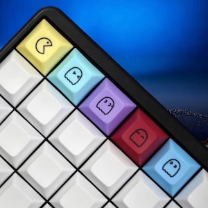 Może przedstawiać: Zbliżenie niestandardowej klawiatury z białymi i kolorowymi nakładkami na klawisze. Nakładki na klawisze przedstawiają wzory Pac-Mana i duchów w kolorze żółtym, niebieskim, fioletowym i czerwonym. Klawiatura ma czarną ramkę i niebieskie tło.